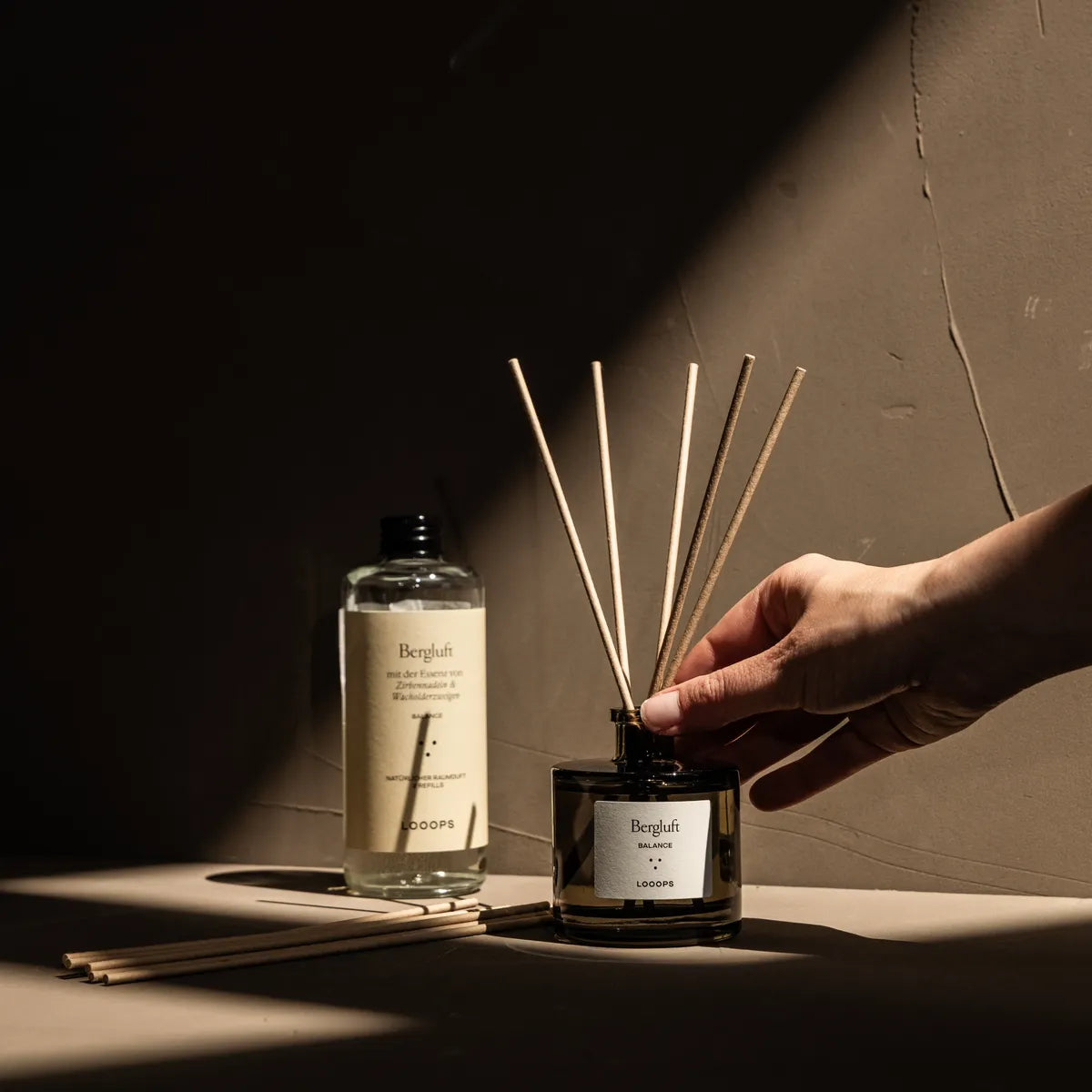 Hand touches stick diffuser and glass bottle in the background