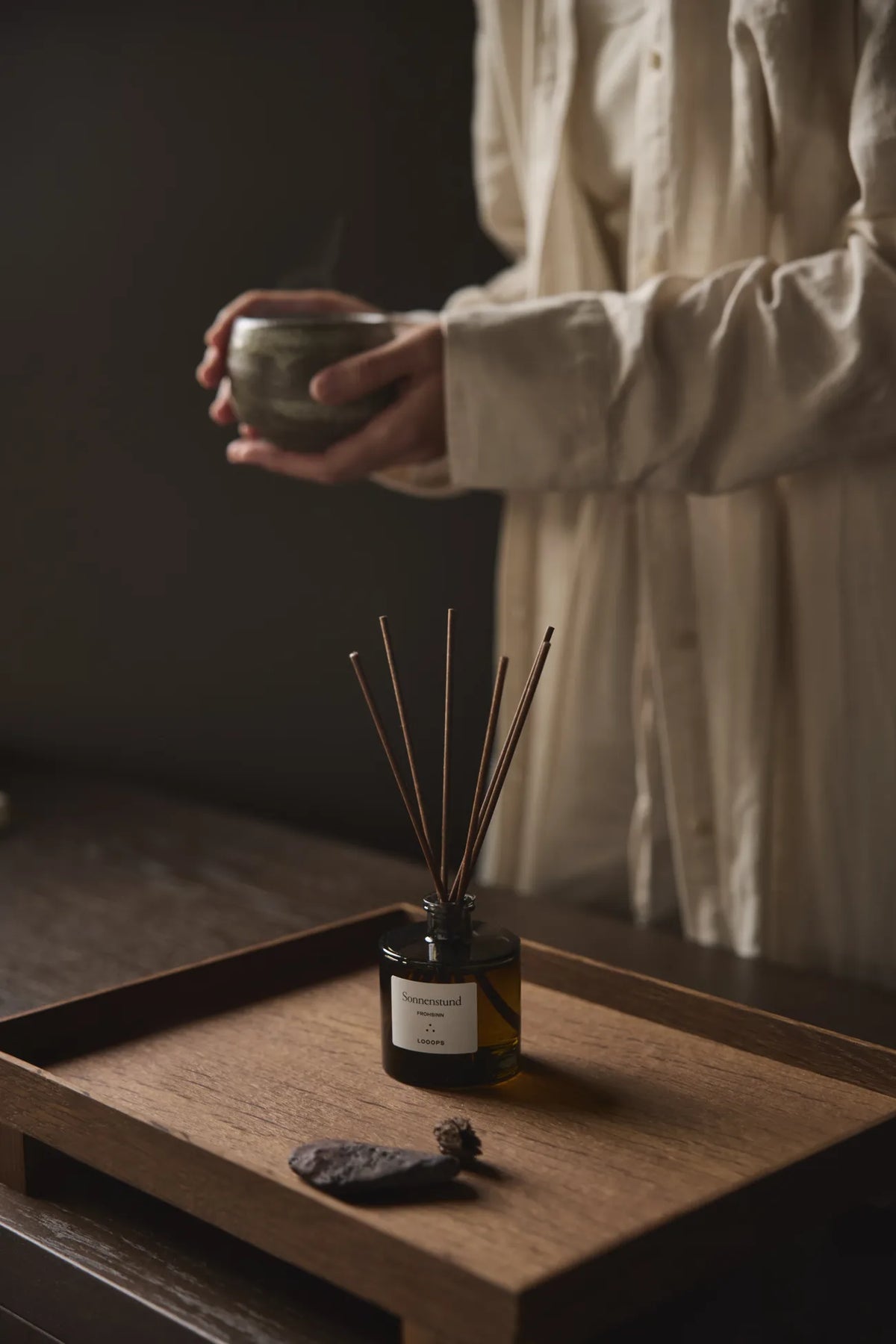 reed diffuser Sonnenstund by Looops on a wooden tray; in the background, a person holding a bowl